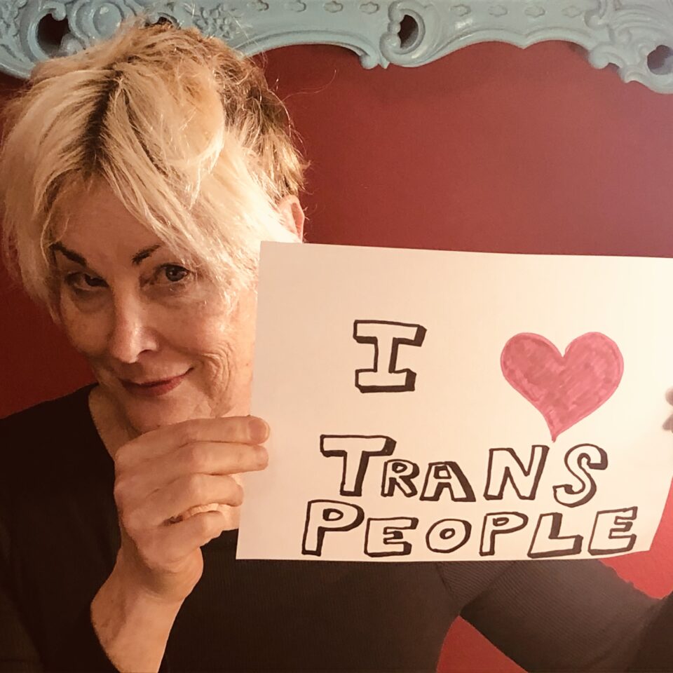 Shawna Virago, a white person with blond hair smiles and holds up a sign that says "I (drawn red heart) Trans People”